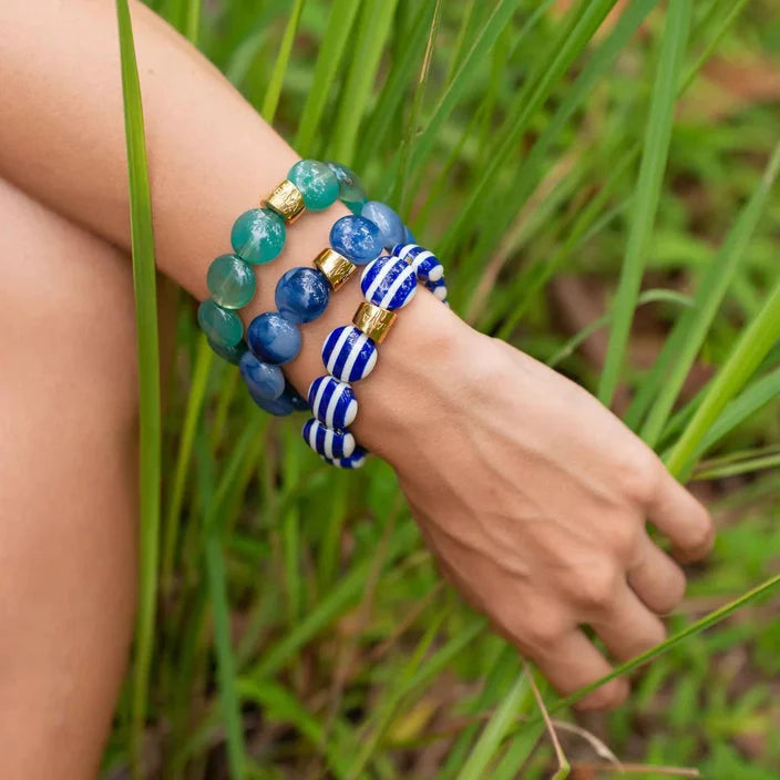 A close-up of a person's wrist wearing three beaded bracelets outdoors among tall green grass. The bracelets include teal and blue round beads, blue and white striped round beads, and blue beads separated by small gold spacer rings.