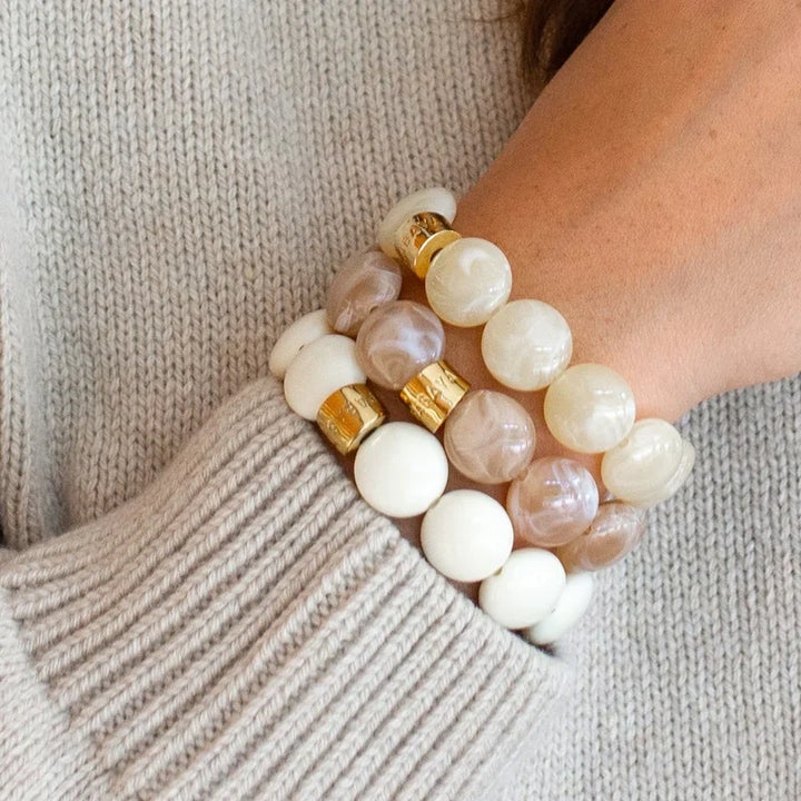 Three beaded bracelets stacked on a person's wrist, featuring smooth white and milky beige beads with two gold-toned cylindrical spacers, shown against a beige knit sweater.