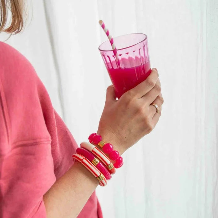 A person in a pink top holds a pink drink in a clear glass with a striped straw, wearing a stack of pink and red beaded bracelets with gold accents on their wrist against a white curtain background.