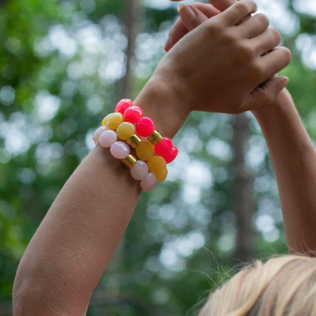 A close-up lifestyle shot of a person's raised arms wearing three beaded stretch bracelets. The bracelets feature glossy round beads in vibrant pink, yellow, and pale peach, separated by small gold metallic spacer rings.