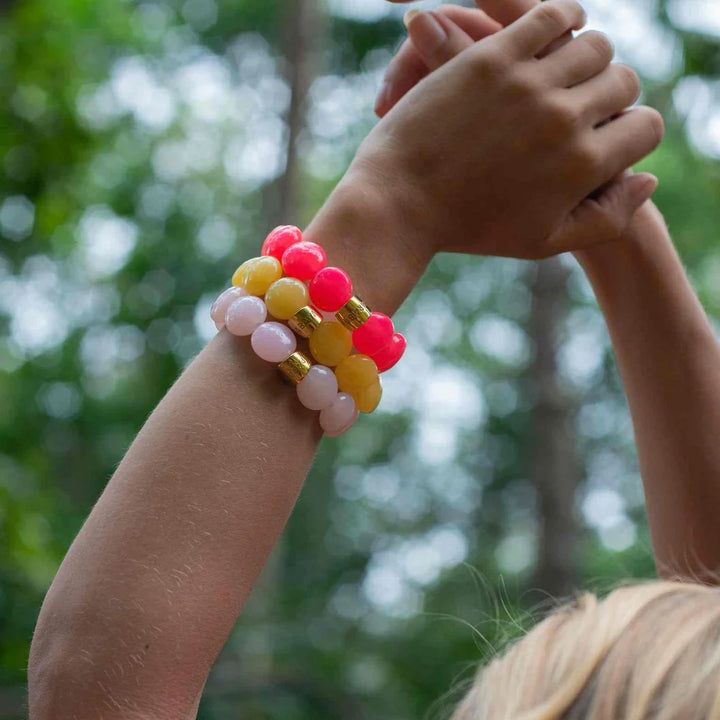 A close-up lifestyle shot of a person's raised arms wearing three beaded stretch bracelets. The bracelets feature glossy round beads in vibrant pink, yellow, and pale peach, separated by small gold metallic spacer rings.