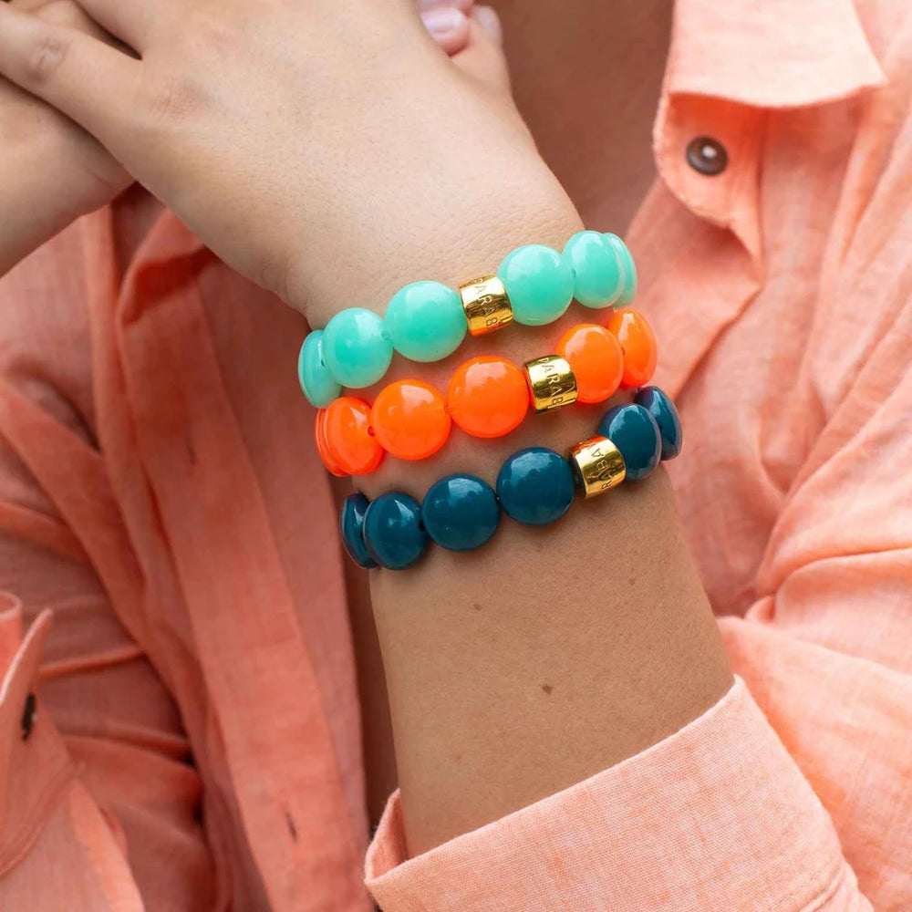Three beaded stretch bracelets in mint green, bright orange, and deep teal, each accented with a small gold spacer, shown stacked on a person's wrist wearing a peach-colored collared shirt.