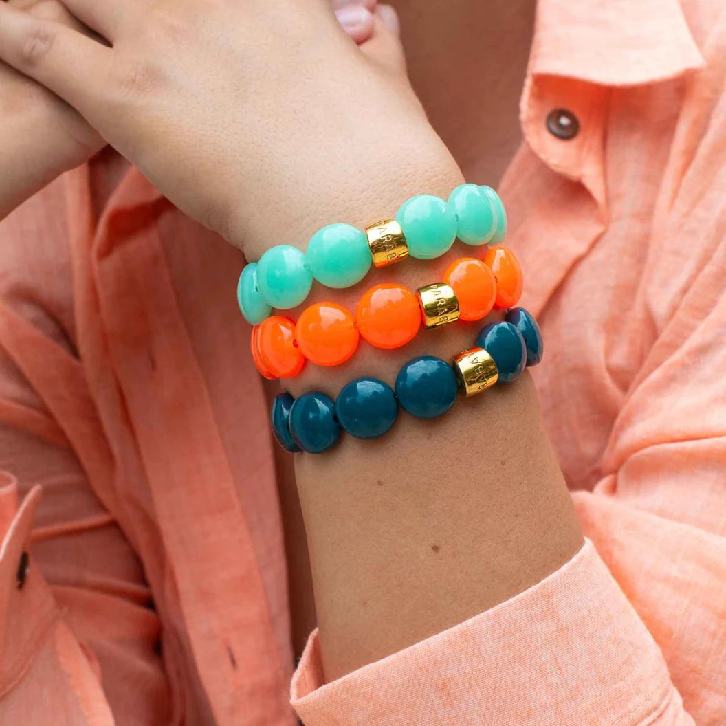 Three beaded stretch bracelets in mint green, bright orange, and deep teal, each accented with a small gold spacer, shown stacked on a person's wrist wearing a peach-colored collared shirt.