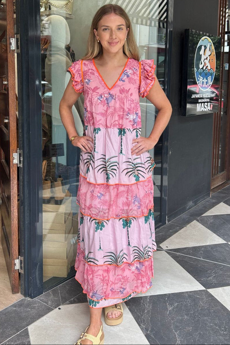 A woman stands in front of a boutique doorway wearing a pink tiered maxi dress with a tropical palm tree print and ruffled cap sleeves. The dress has a floral pink upper section and a lower section with black palm tree silhouettes on a white background.