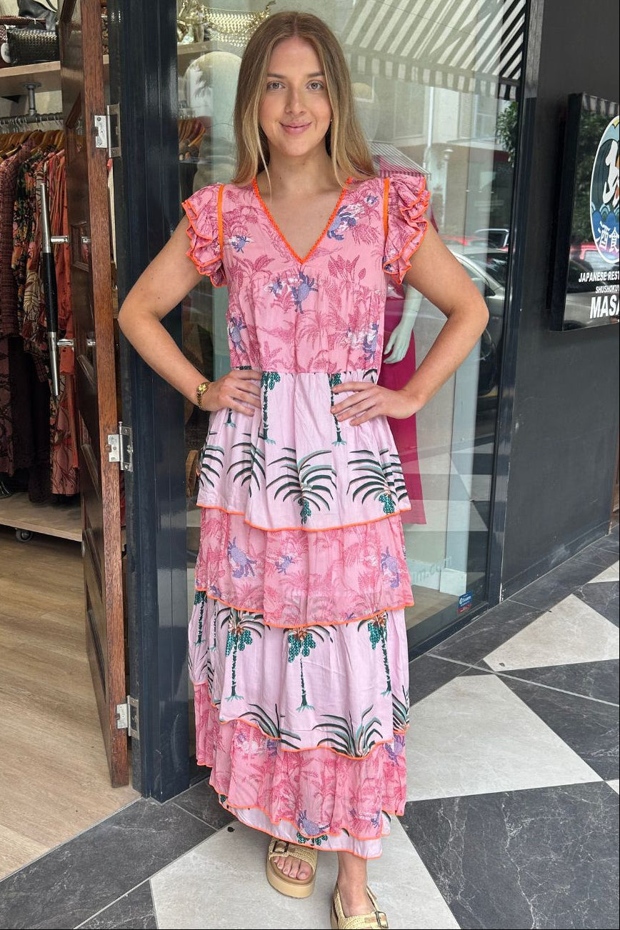 A woman modeling a pink tiered maxi dress with a tropical palm tree print, standing in front of a boutique storefront.