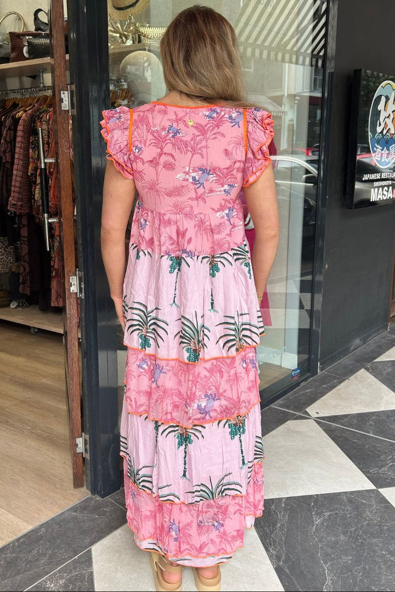 A woman viewed from behind wearing a pink tiered maxi dress with a tropical palm tree print, standing in front of a boutique storefront.