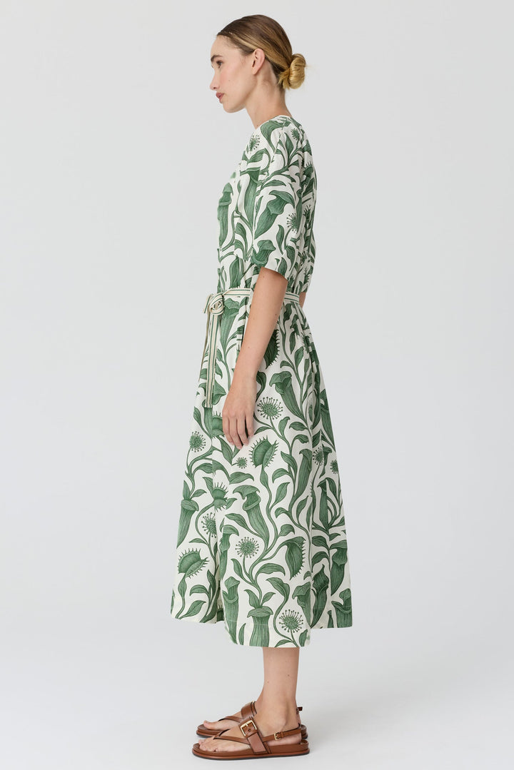 A woman in profile wearing a green and white tropical leaf print midi dress with a tie waist belt, paired with brown leather sandals, against a light gray studio background.