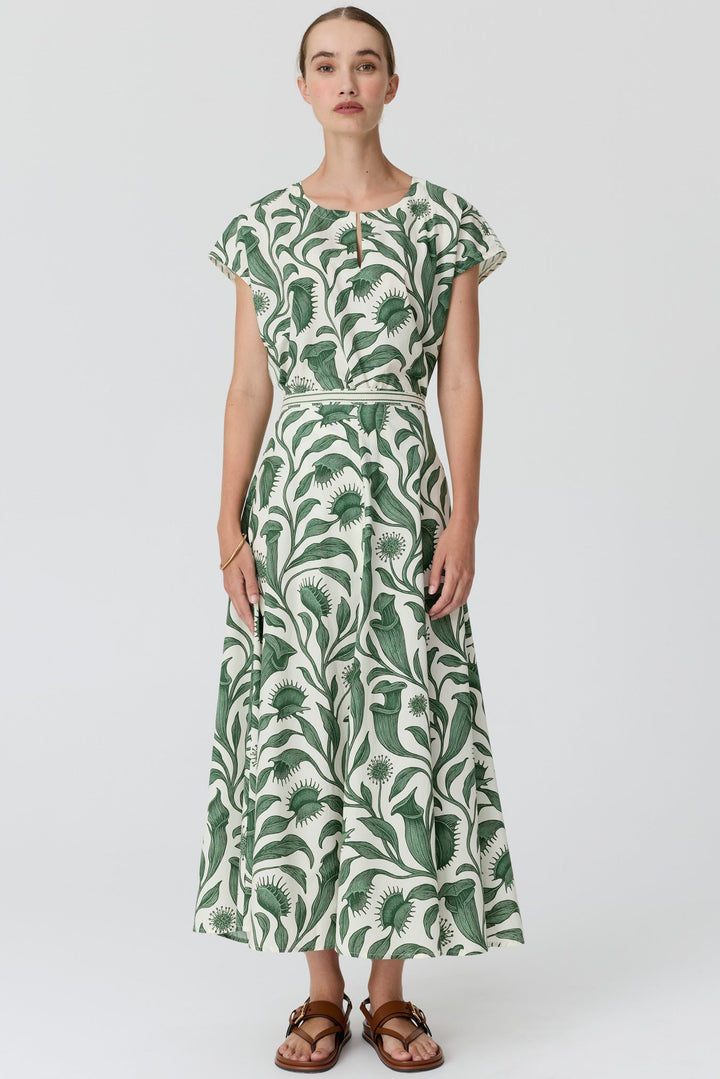 A woman modeling a long-sleeved maxi dress with a green and white botanical leaf pattern, standing against a light gray studio background.