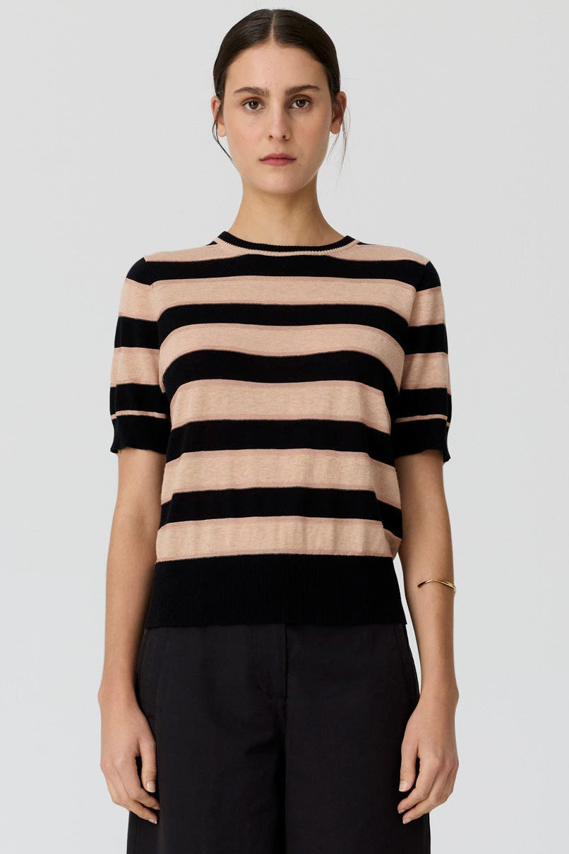 A model stands against a light gray studio background wearing a short-sleeve knit top with horizontal beige and black stripes, black wide-leg pants, and black sandals.