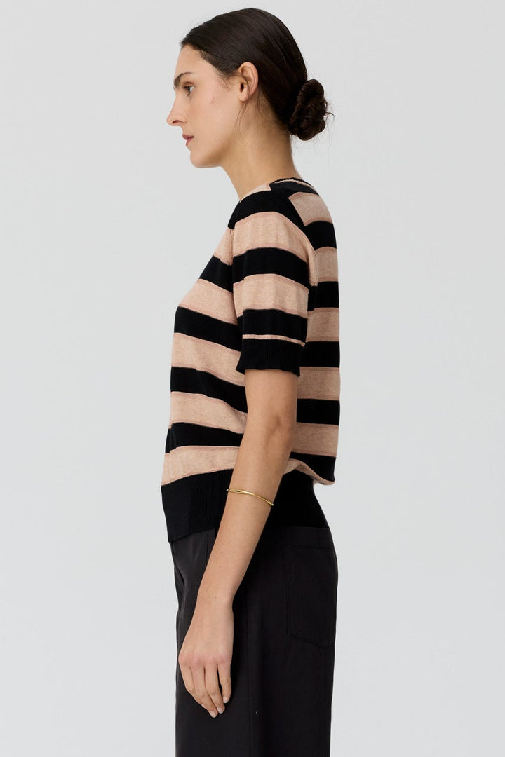 A woman stands in profile wearing a short-sleeve knit top with horizontal black and beige stripes, paired with black wide-leg trousers and black flip-flops, against a light gray studio background.