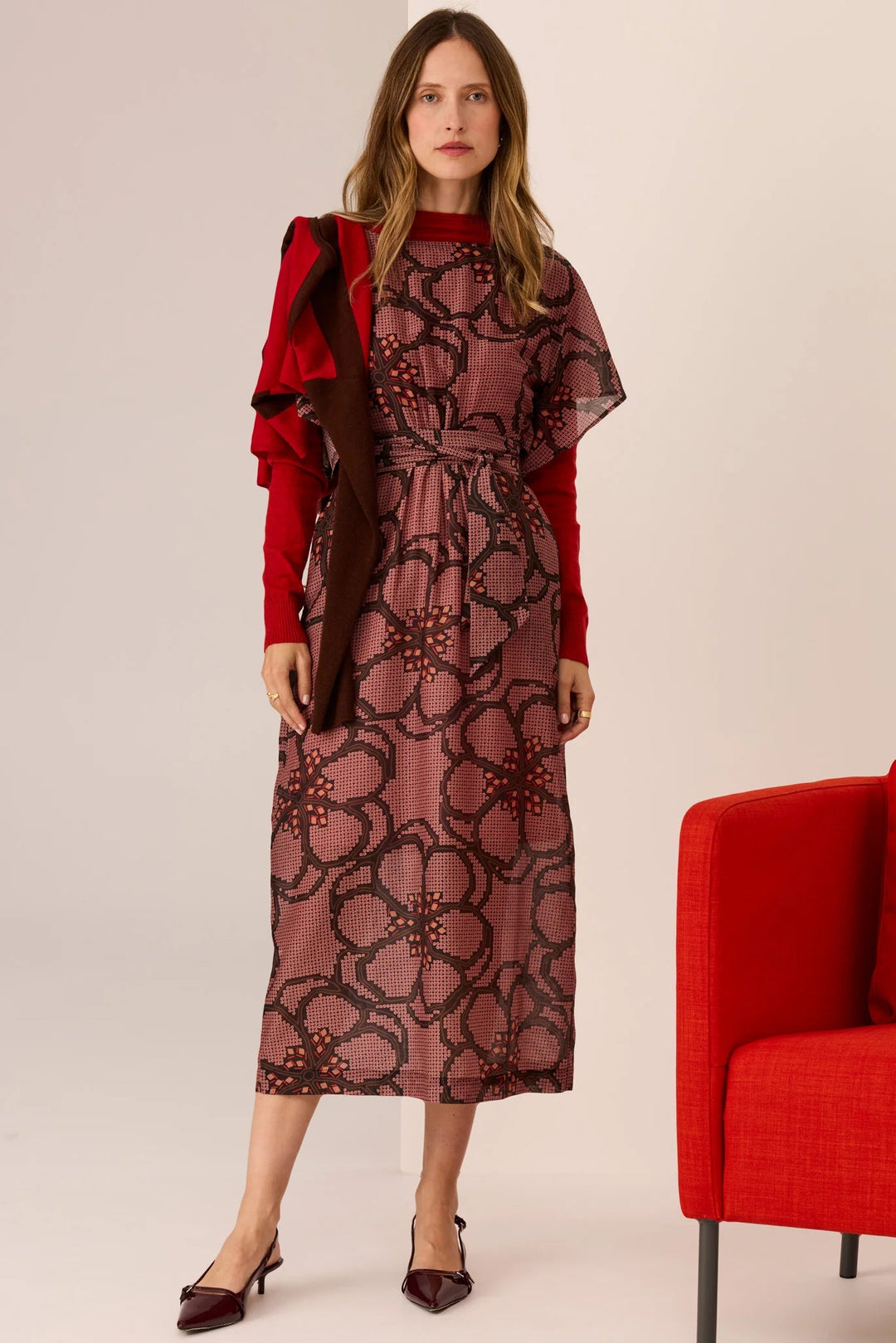 Woman wearing a patterned dress with a red jacket and shoes, standing next to a red chair on a plain background.
