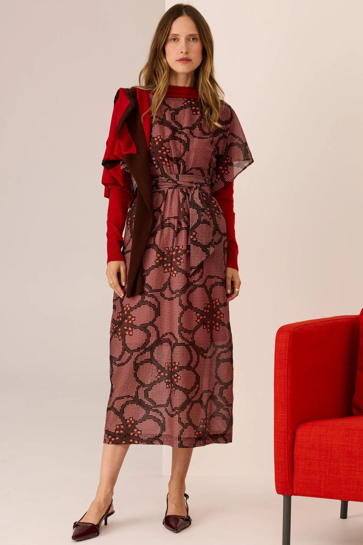 Woman wearing a patterned dress with a red jacket and shoes, standing next to a red chair on a plain background.