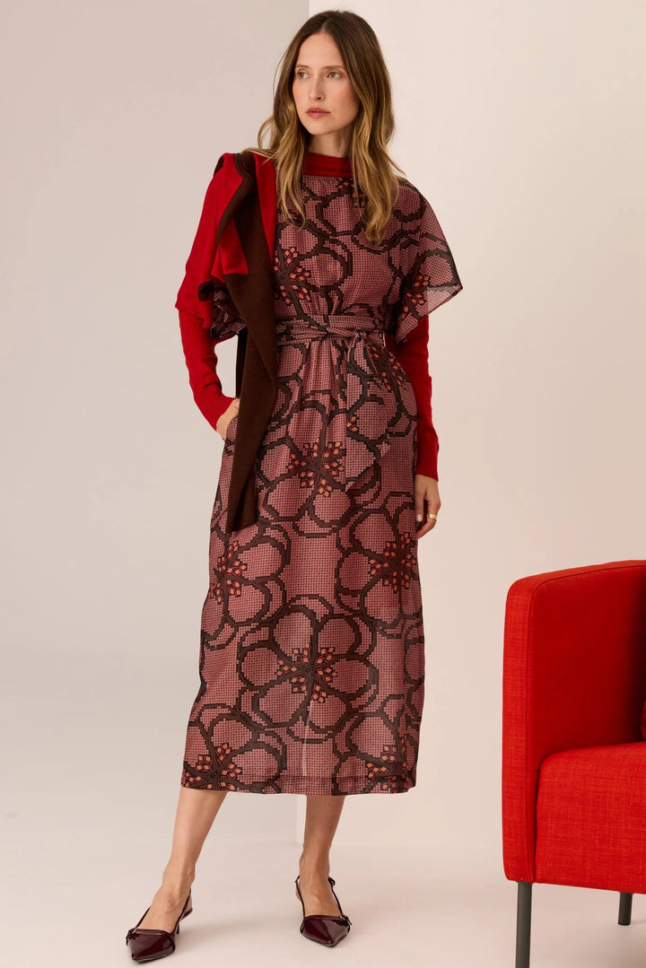 Woman wearing a patterned dress with a red jacket and shoes, standing next to a red chair on a beige background