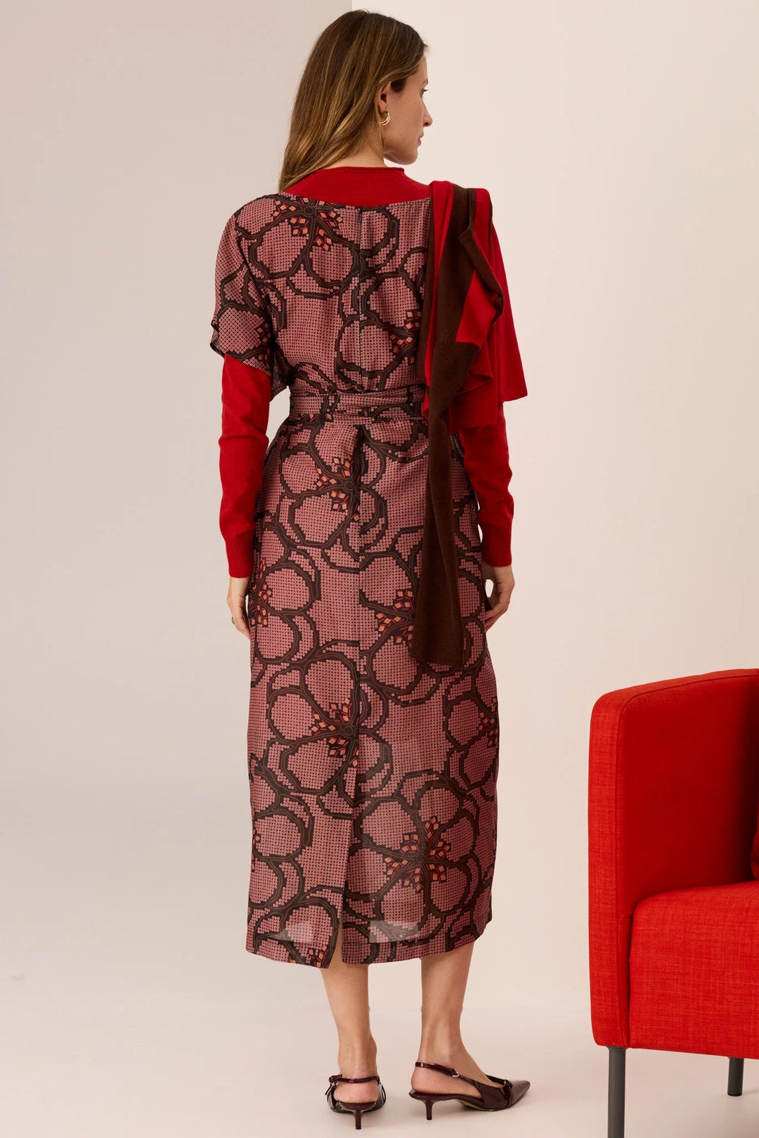 Woman wearing a red and patterned dress standing next to a red chair on a plain background