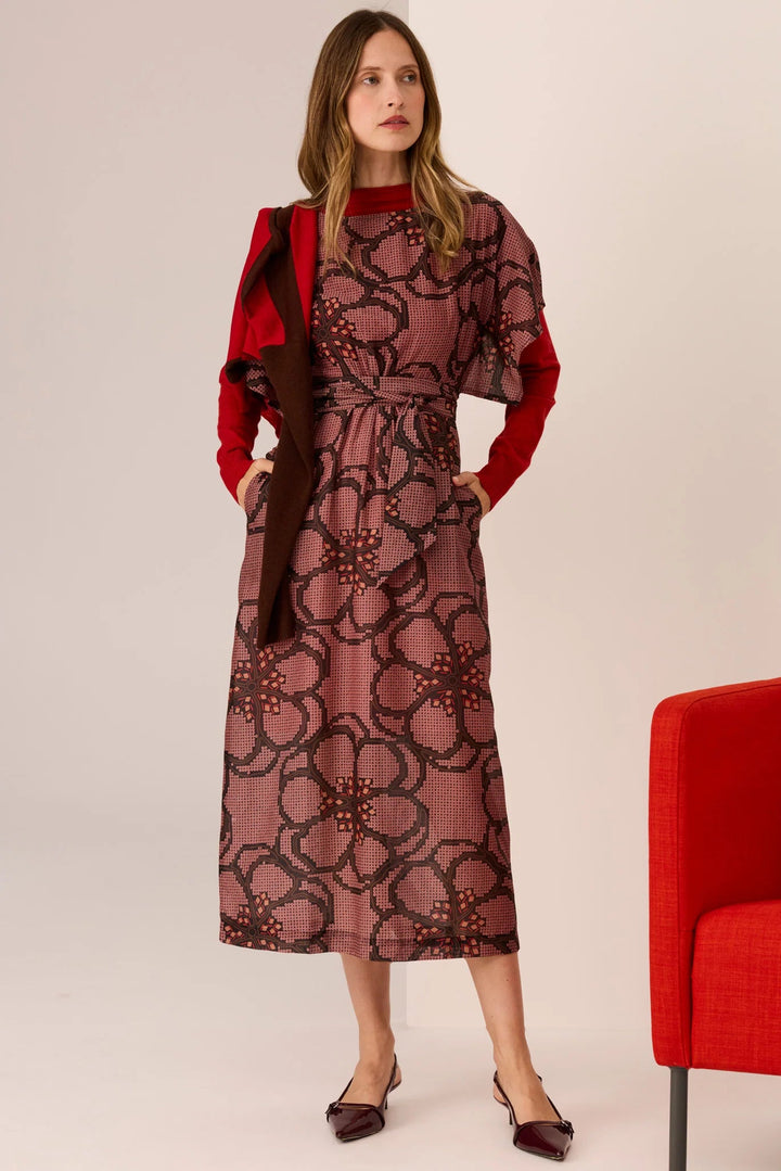 Woman wearing a patterned dress with a red jacket and shoes, standing next to a red chair on a plain background.