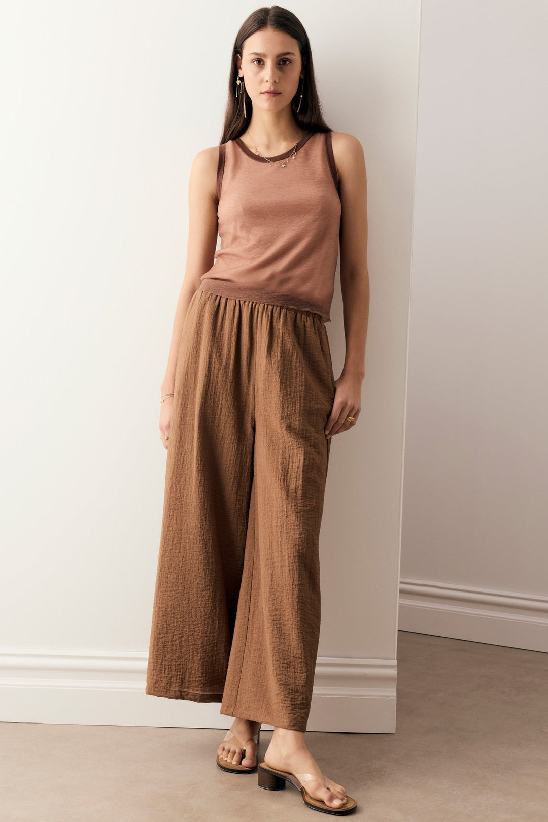 A woman modeling a sleeveless dusty rose top and matching brown wide-leg pants, standing in a minimalist room with white walls and a beige floor.
