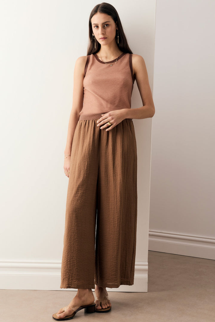A woman modeling a sleeveless dusty rose top and matching brown wide-leg pants in a minimalist indoor setting with white walls and a beige floor.