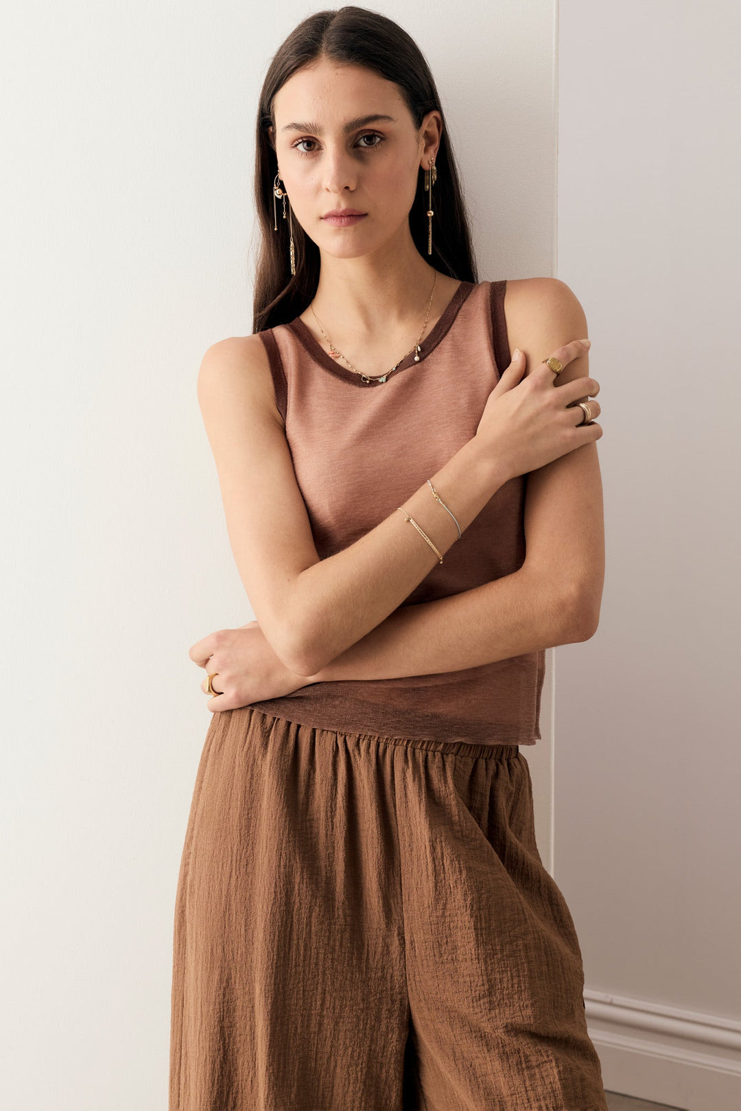 A woman modeling a sleeveless dusty rose top and brown wide-leg pants, accessorized with gold jewelry, standing against a plain off-white wall.