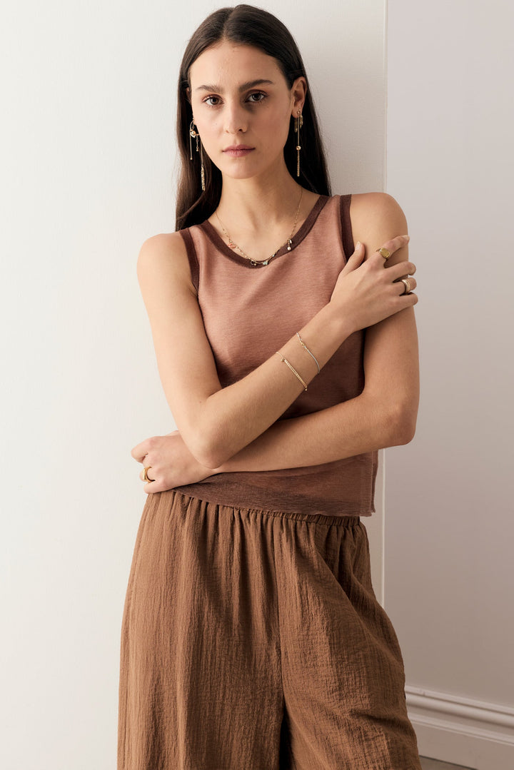 A woman modeling a sleeveless dusty rose top and brown wide-leg pants, accessorized with gold jewelry, standing against a plain off-white wall.