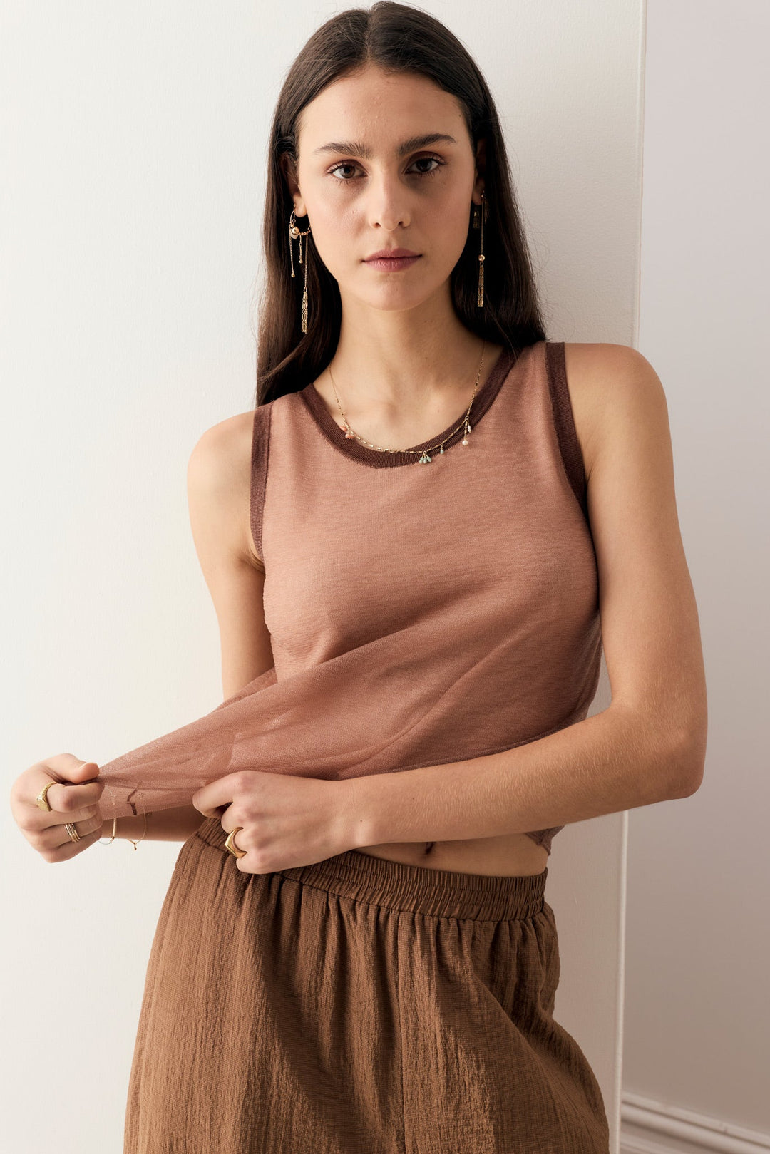 A woman modeling a sleeveless dusty rose-brown tank top with dark brown trim and a matching brown skirt against a plain off-white wall.