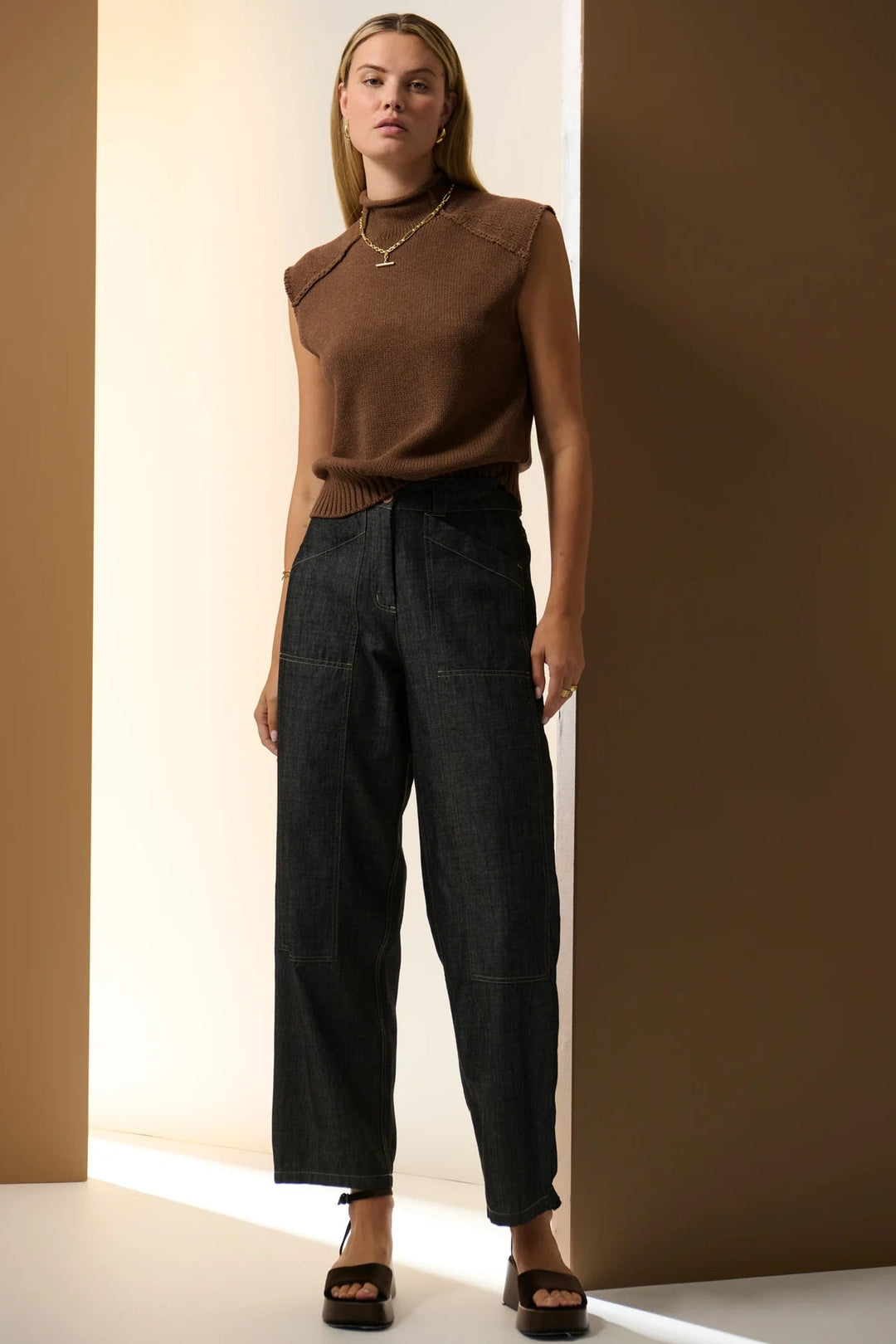 Woman wearing a brown top and dark jeans standing against a neutral wall. Pol Muhle Barrel Denim Pant Indigo