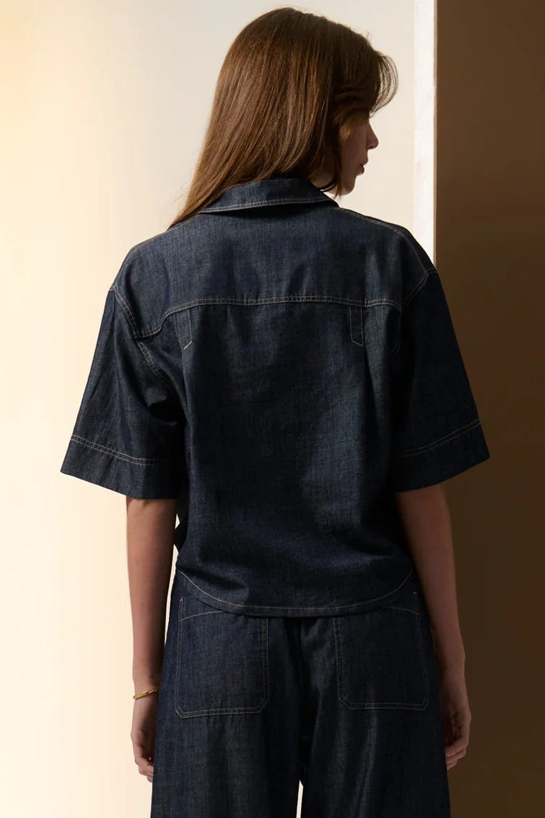 Person wearing a dark denim jumpsuit standing against a plain background. Pol Muhle Denim Top Indigo