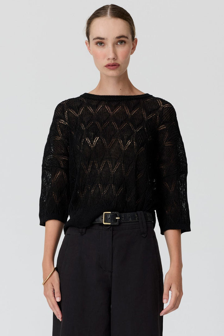 A model stands against a light gray studio background wearing a black three-quarter sleeve top with a textured openwork pattern, paired with black wide-leg trousers and brown sandals.