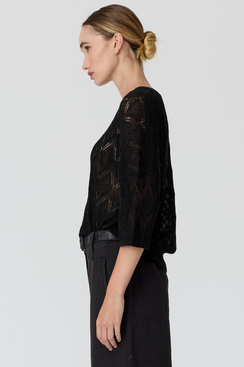 A woman stands in profile wearing a black sheer lace top with elbow-length sleeves and black wide-leg trousers, photographed against a clean light gray studio background.