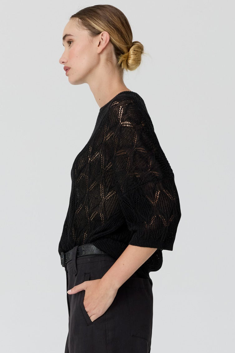 A woman stands in profile wearing a black sheer lace top with elbow-length sleeves and black wide-leg trousers, photographed against a light gray studio background.