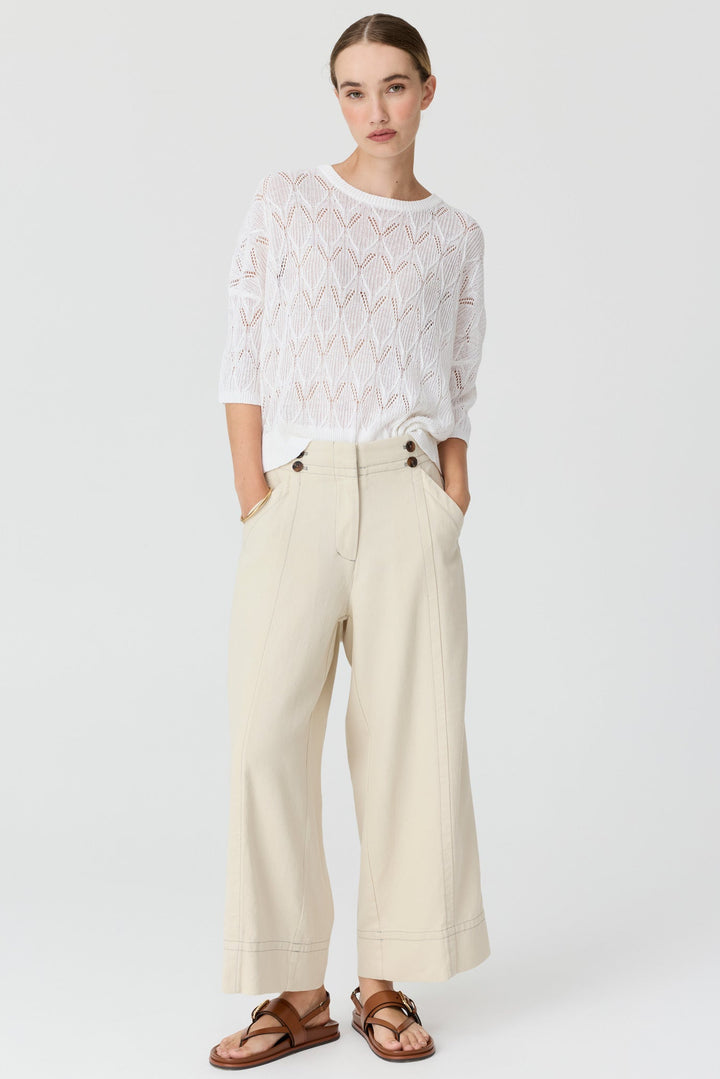 A model stands against a light gray studio background wearing a white textured lace long-sleeve top, cream wide-leg cropped pants, and brown leather sandals.