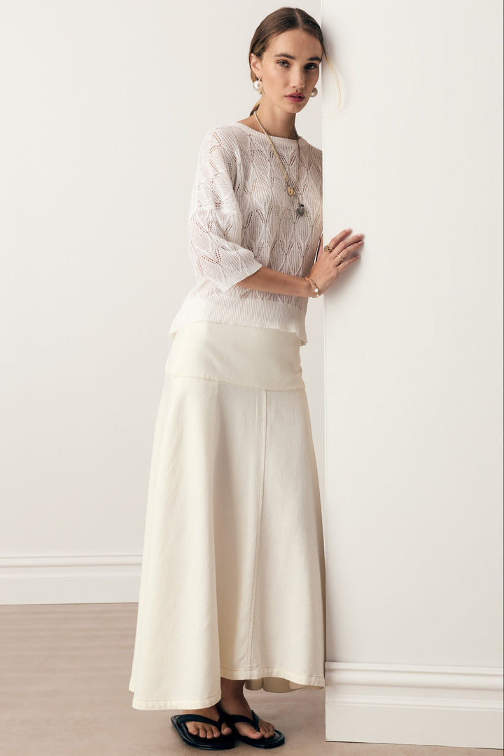 A woman stands against a white wall wearing a cream-colored two-piece outfit consisting of a sheer lace long-sleeve top and a matching high-waisted maxi skirt, paired with black sandals.
