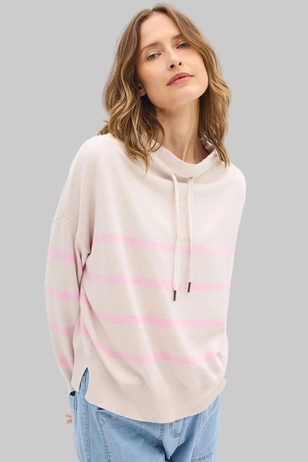 Woman wearing a beige sweater with pink stripes and blue jeans on a white background