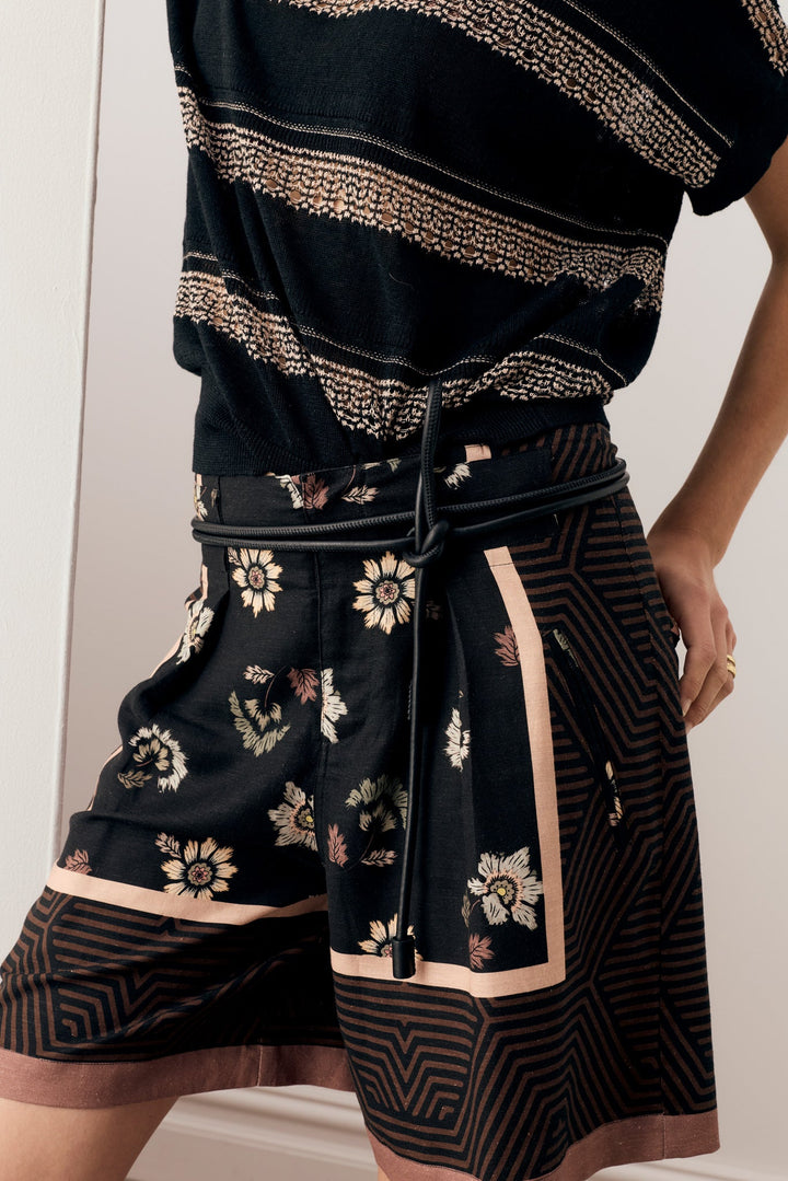 A close-up of a person wearing a black patterned short-sleeve top and matching black shorts with beige and white floral motifs, a drawstring waist, and geometric stripe accents.