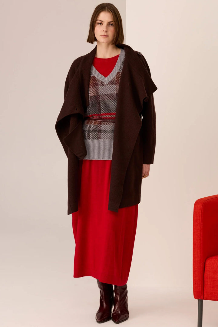 Woman wearing a brown coat over a red dress with a patterned top, standing against a plain background.