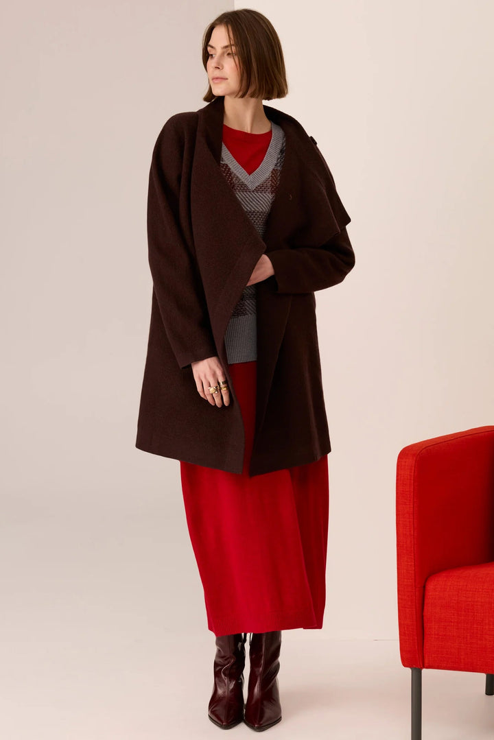 Woman wearing a brown coat over a red dress with a gray sweater, standing against a plain background.