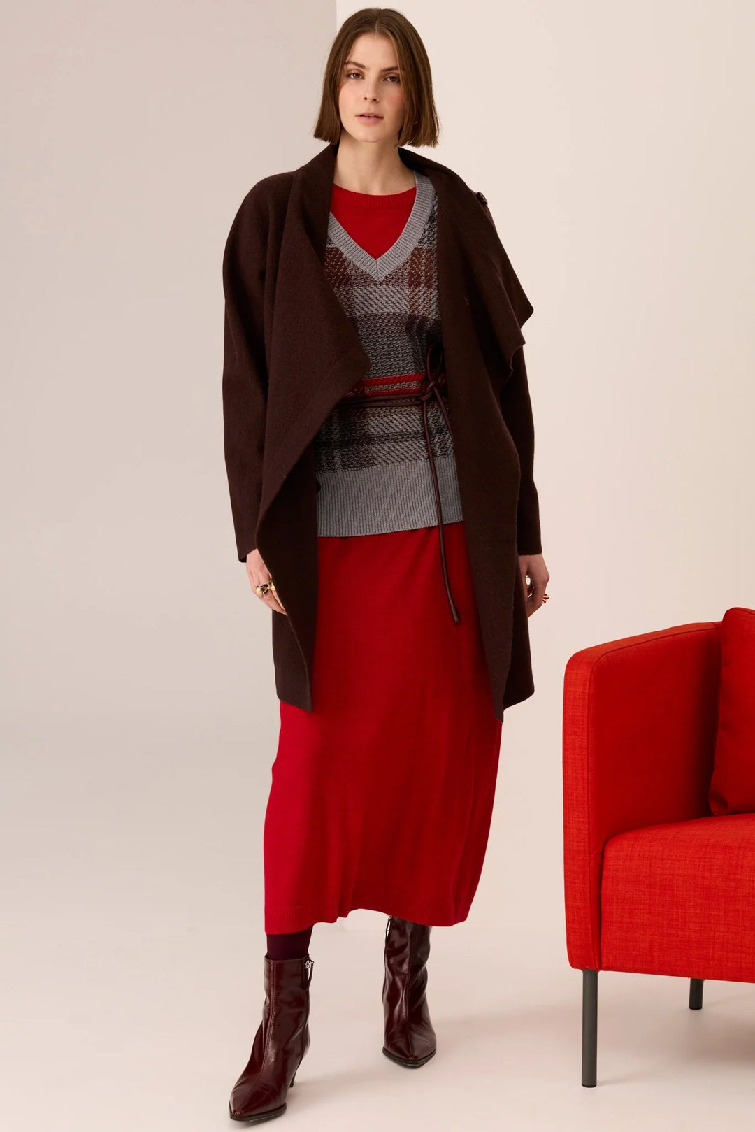 Woman wearing a red dress with a brown coat and boots, standing next to a red chair on a plain background