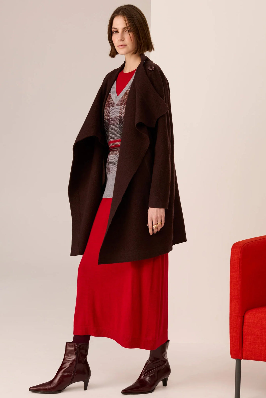 Woman wearing a brown coat over a red dress with a gray sweater, standing next to a red chair on a plain background.