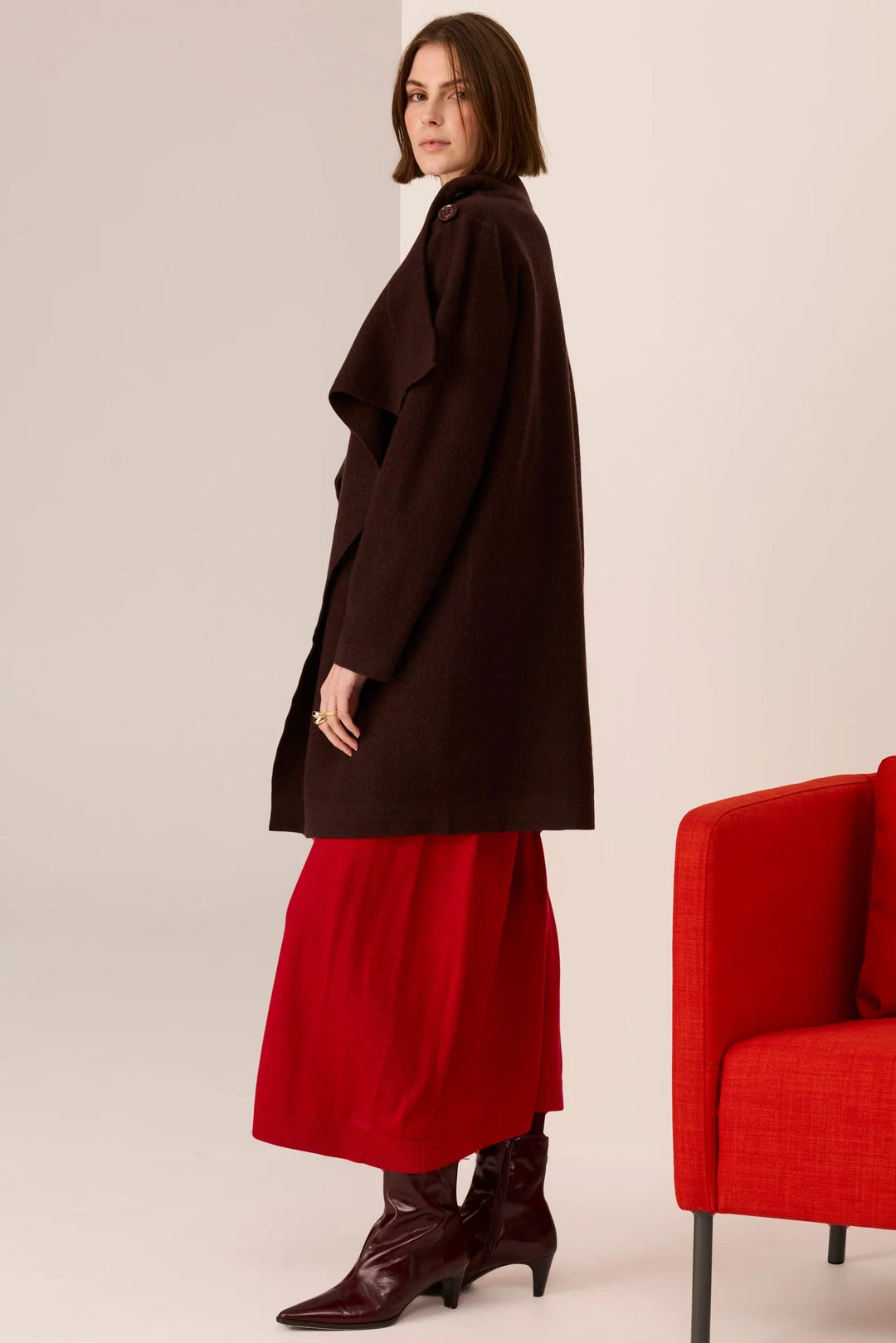 Woman wearing a brown coat and red skirt standing next to a red chair on a plain background