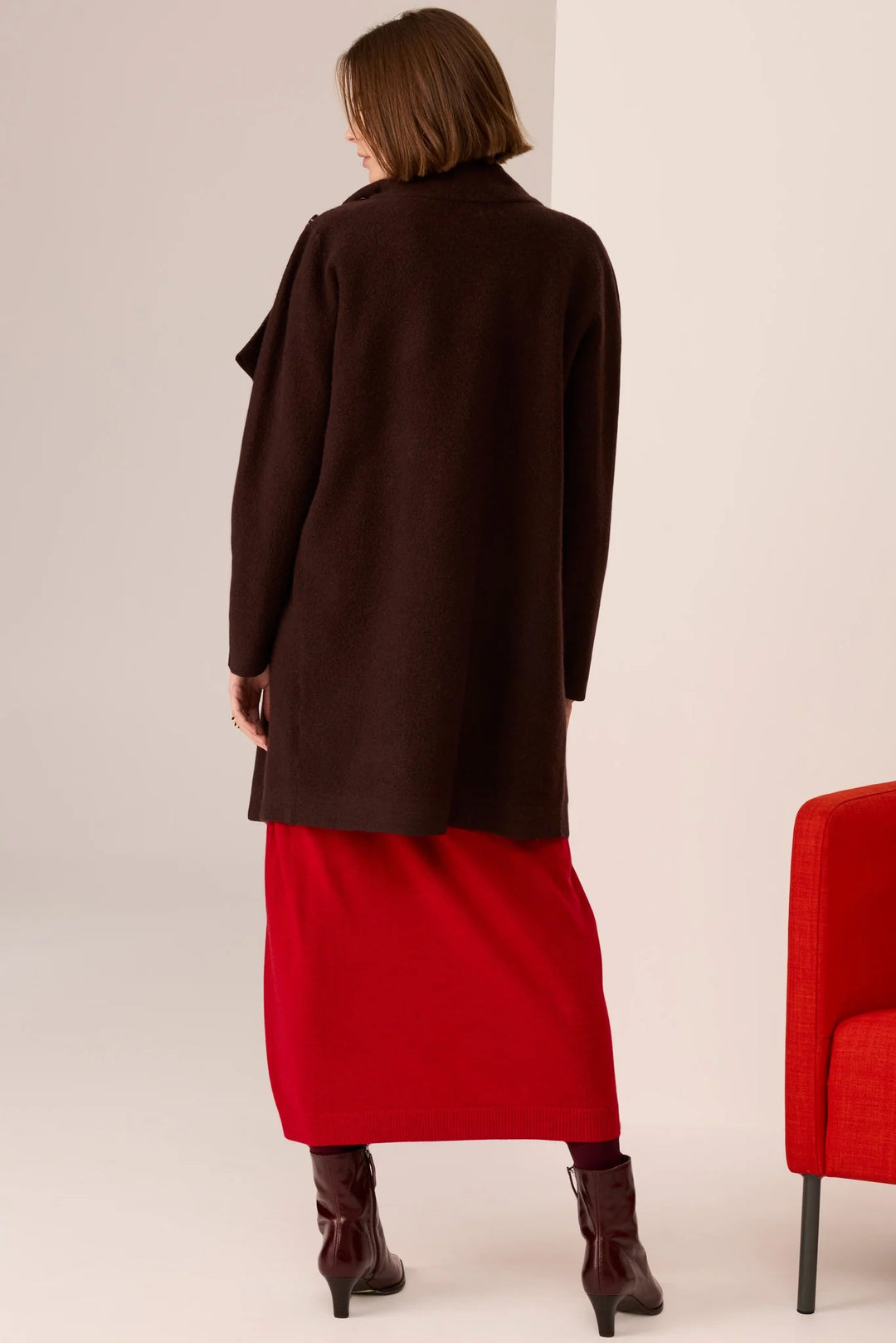 Person wearing a long brown coat over a red dress, standing next to a red chair on a plain background