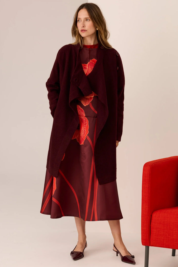 Woman wearing a dark red outfit with red accents standing next to a red chair on a plain background