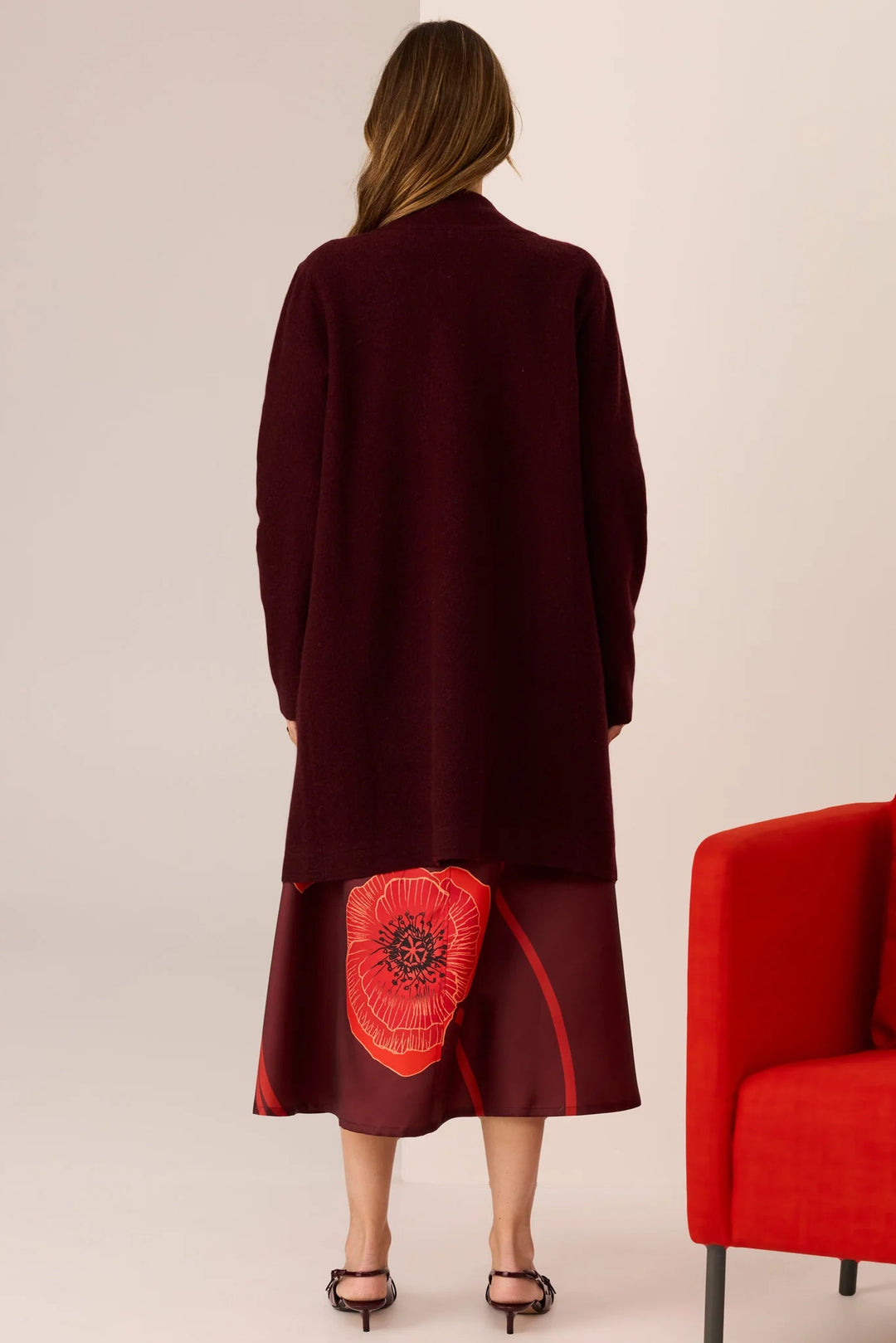 Woman wearing a burgundy coat with a red floral design on the back, standing next to a red chair.