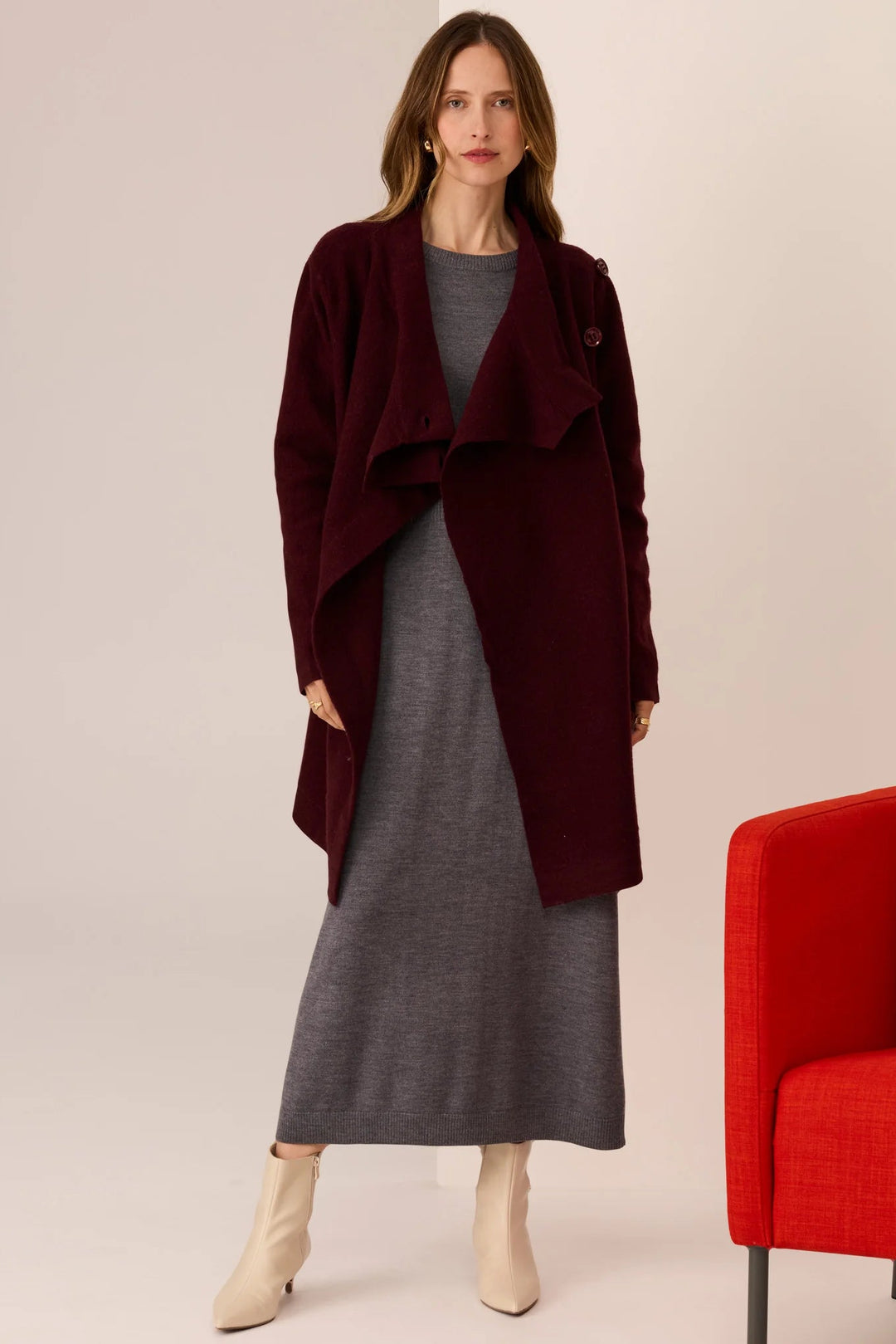 Woman wearing a burgundy coat over a gray dress with a red chair in the background