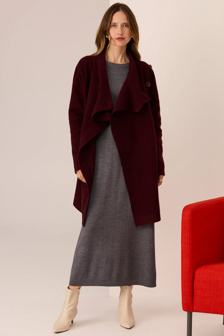 Woman wearing a burgundy coat over a gray dress with a red chair in the background