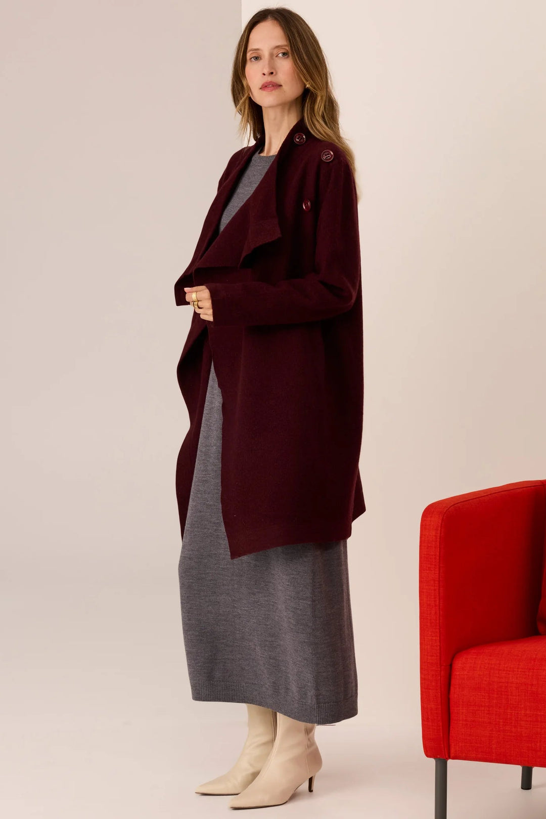 Woman wearing a long burgundy coat over a gray dress, standing next to a red chair on a beige background