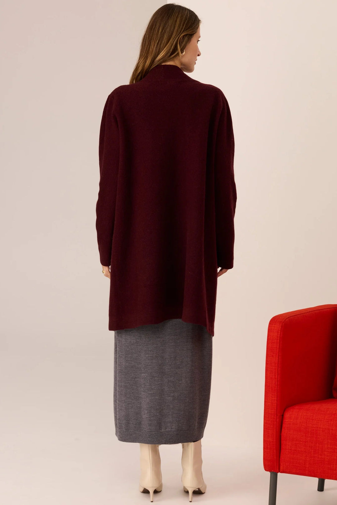 Woman wearing a burgundy coat standing next to a red chair on a beige background