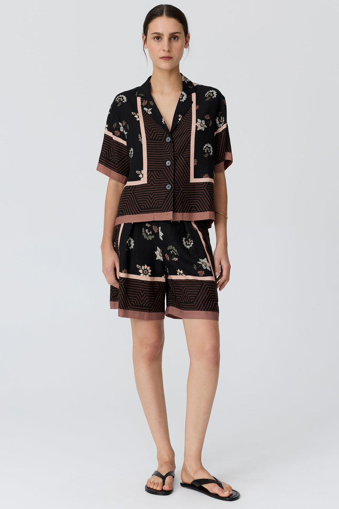 A woman modeling a matching short-sleeve button-front top and shorts set in black with beige and brown geometric patterns and floral accents, paired with black flip-flops against a light gray studio background.