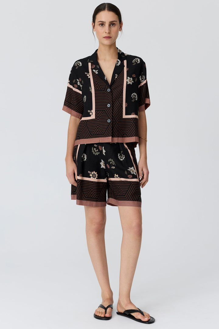 A woman modeling a matching short-sleeve button-front top and shorts set in black with beige and brown geometric patterns and floral accents, paired with black flip-flops against a light gray studio background.