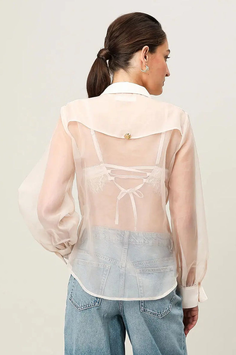 Woman wearing a sheer beige blouse and denim shorts on a plain background