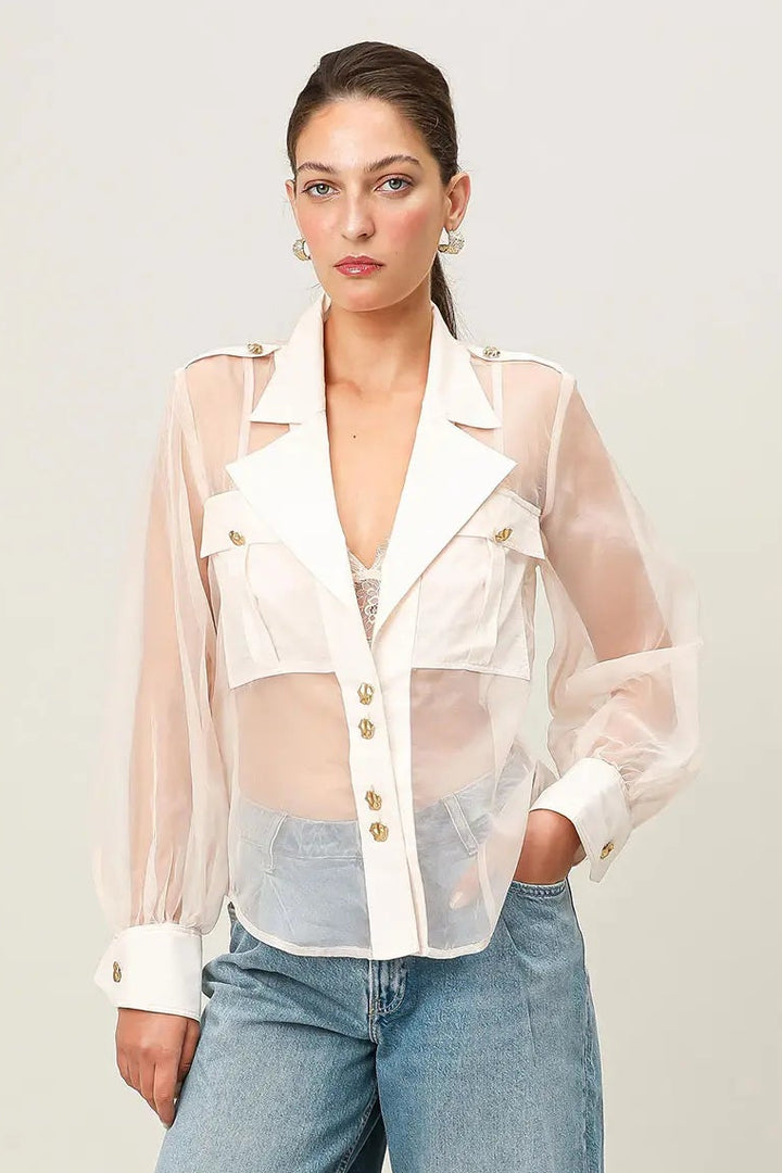 Woman wearing a sheer white blouse over a white shirt with blue jeans on a light gray background