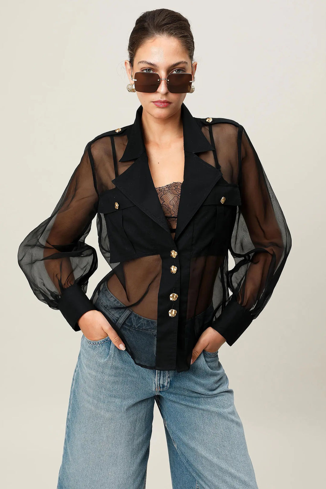Woman wearing a black sheer jacket with gold buttons and blue jeans on a plain background